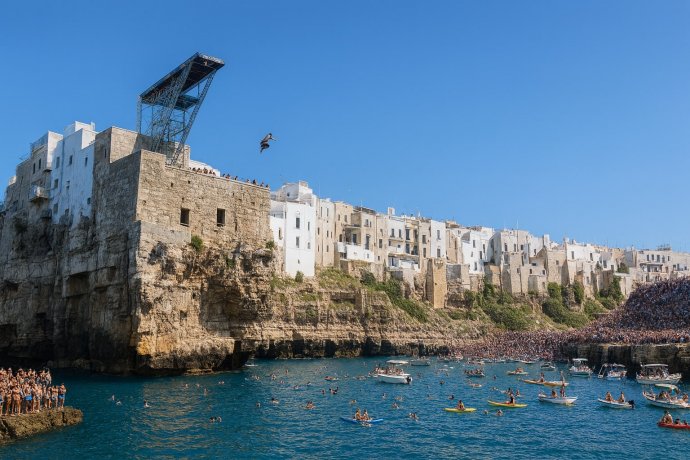 Atleta in volo dal trampolino a Polignano a Mare, folla e barche sotto le scogliere.