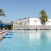 Piscina con sedie a sdraio e ombrelloni blu, edificio bianco e palme sullo sfondo.